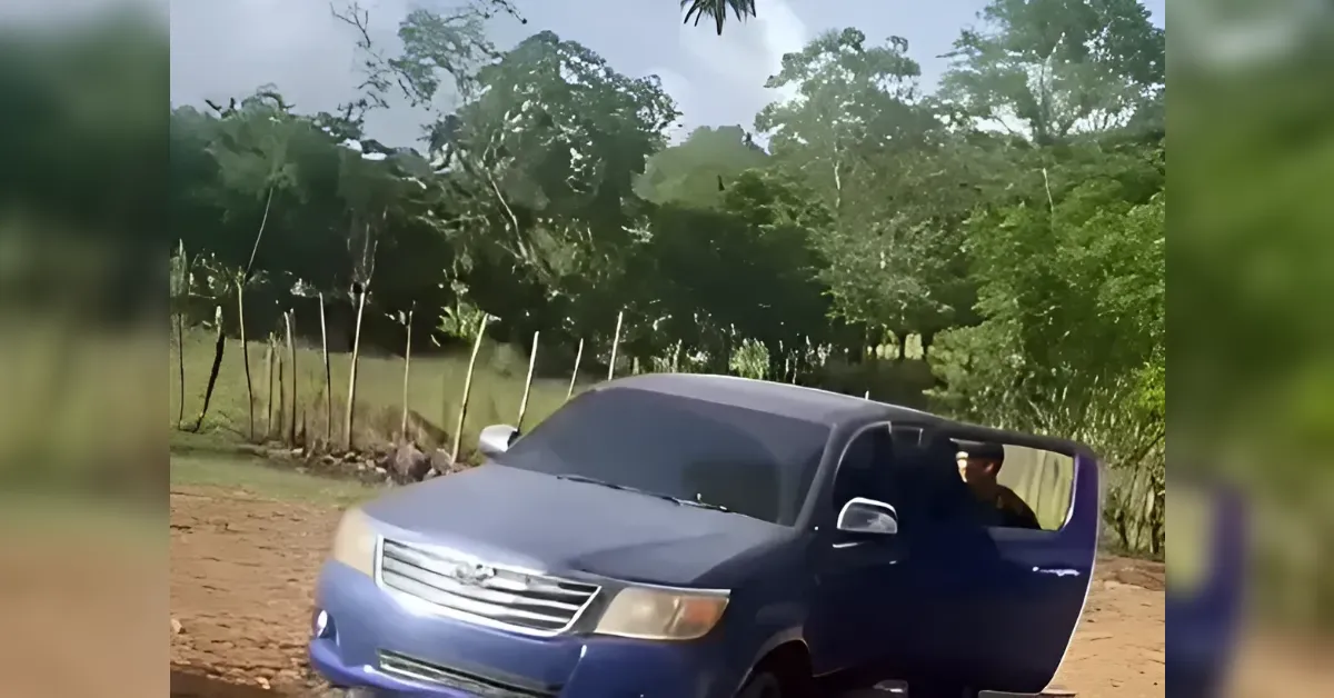 Blue SUV on a dirt road with the driver's side door open and a person partially inside the vehicle amid a rural, tree-filled background.