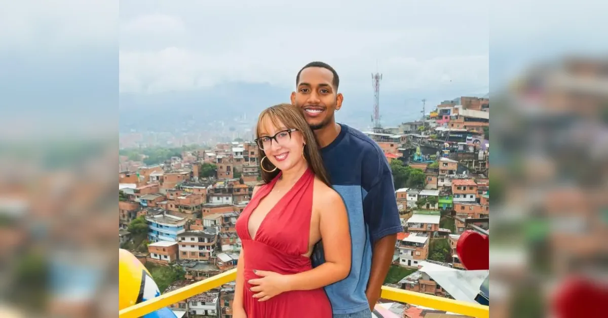 Couple smiling and embracing on a balcony with a densely built hillside city in the background; woman wears a red halter dress, man in a blue T-shirt nearby.