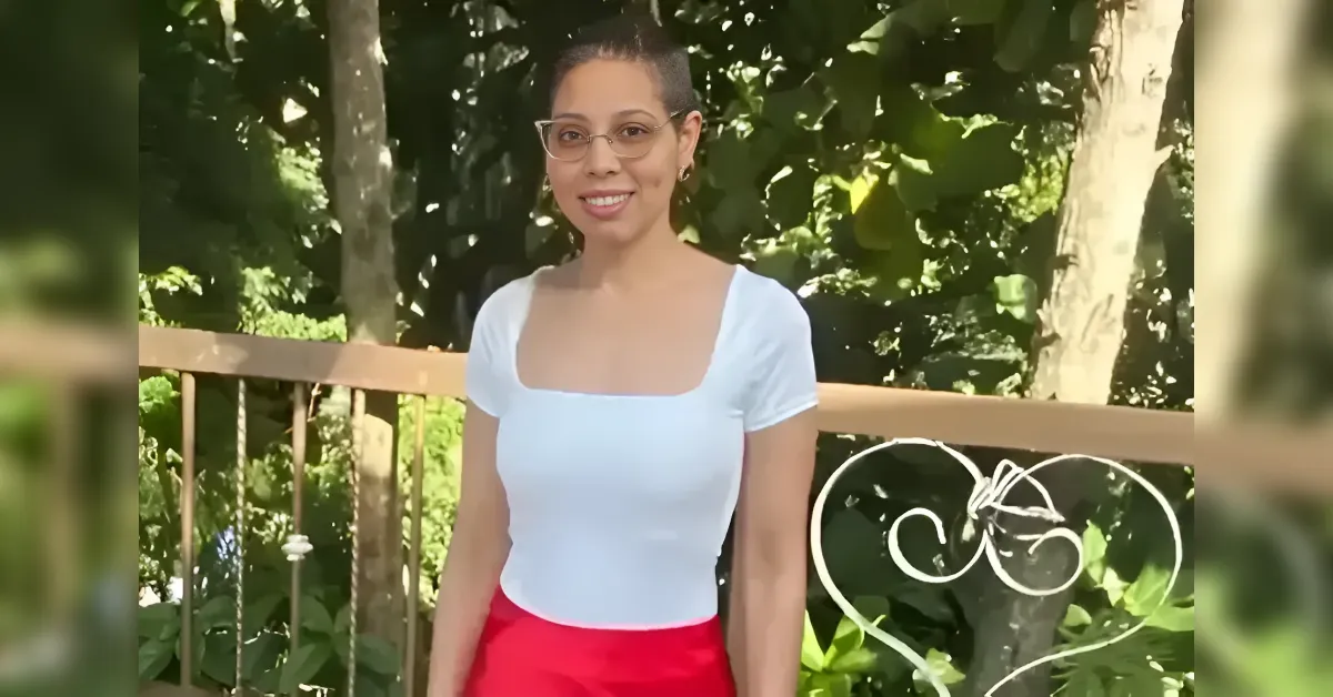 Woman in a white square-neck top and red skirt standing on a wooden balcony railing with green trees in the background, smiling outdoors in daylight.