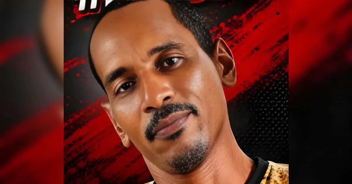 Close-up portrait of a man with short black hair and a styled goatee, looking at the camera against a red and black abstract background.
