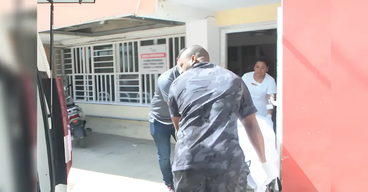 Man in a camouflage shirt walks toward the doorway of a small building; a person in gray stands behind him near the entrance where a woman in white stands inside.