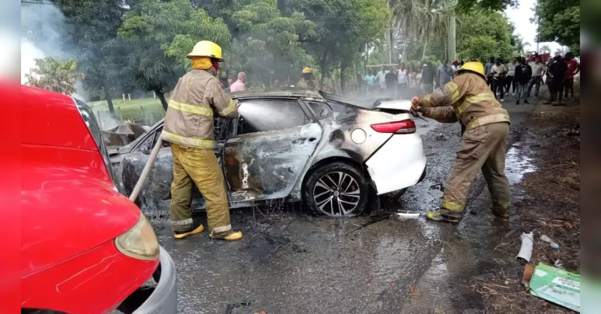 Firefighters in yellow helmets extinguish a charred white sedan after a car fire, with onlookers in the background.