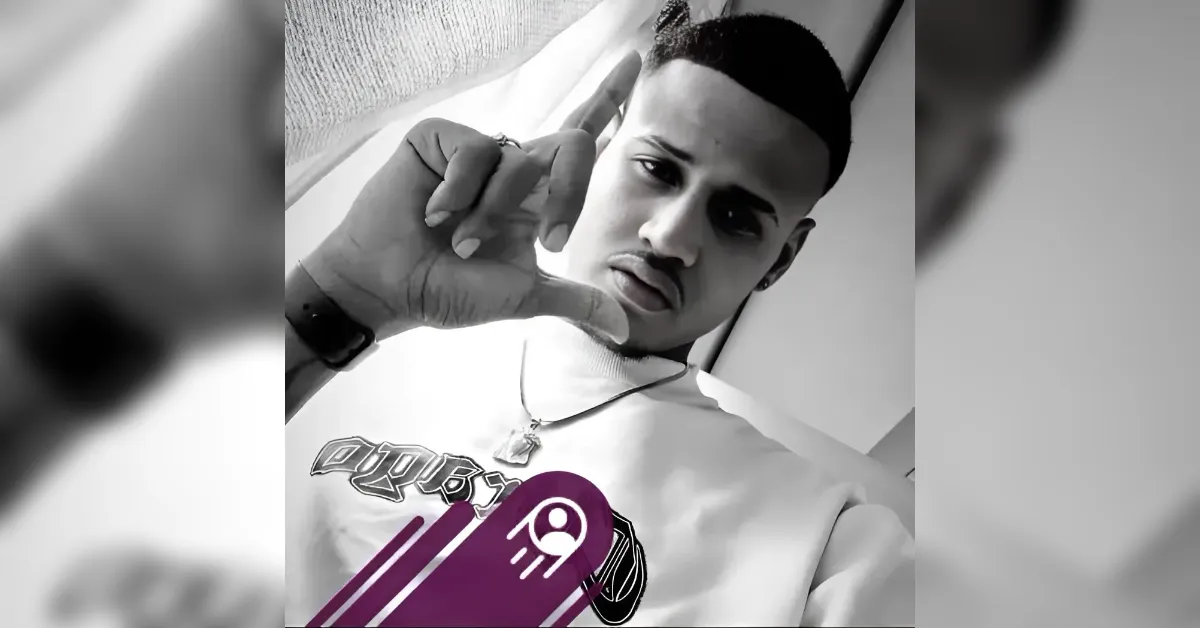 Black-and-white photo of a young man taking a selfie, making a thumbs-down gesture toward the camera, wearing a white graphic tee and a necklace.