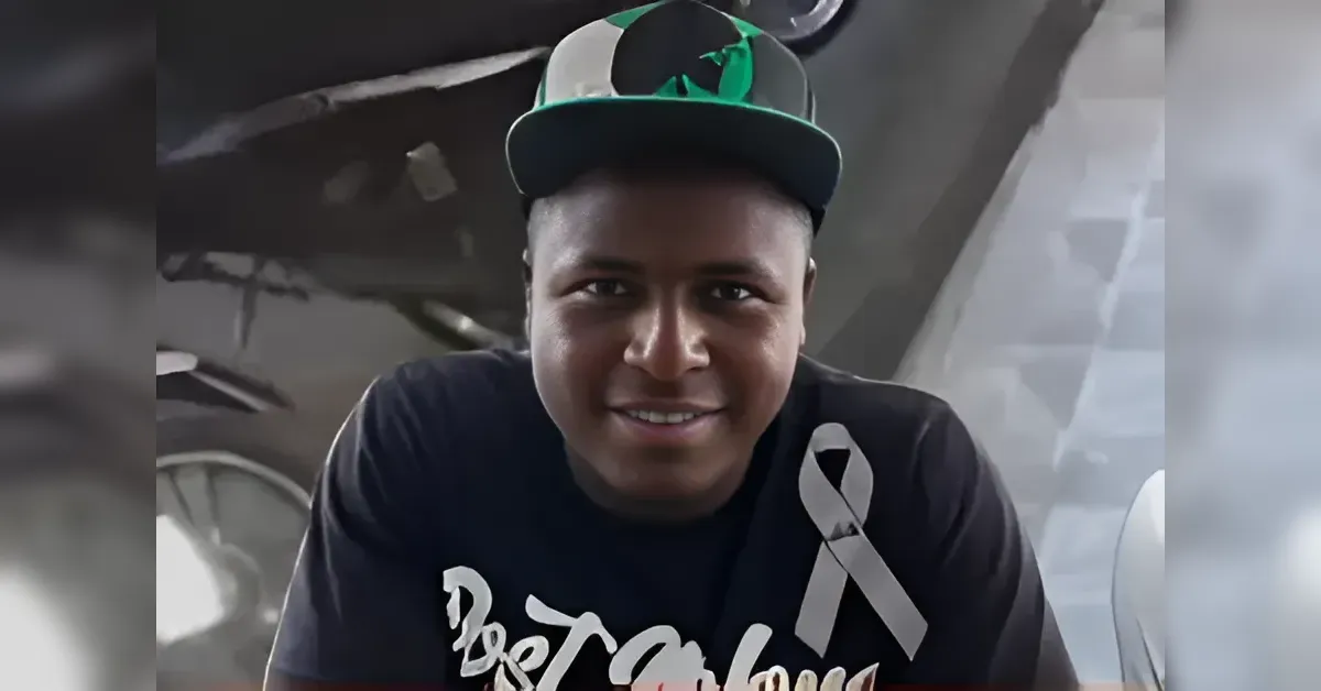 Smiling man wearing a dark T‑shirt with a white awareness ribbon and a green-and-gray cap, looking at the camera.