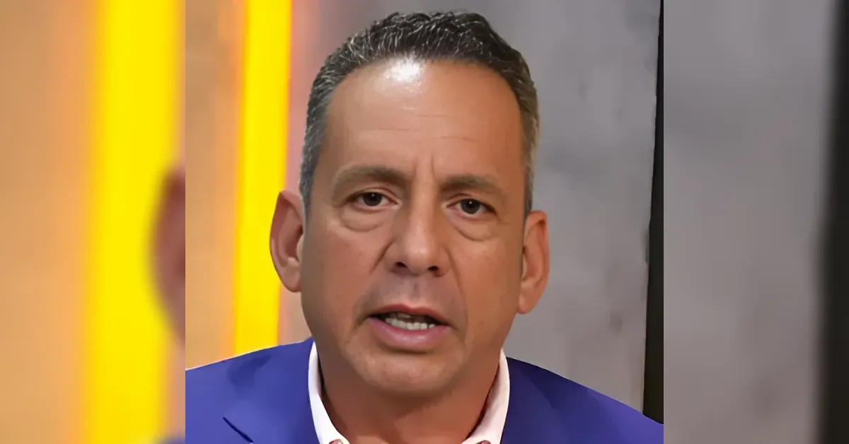 Close-up of a man in a blue suit speaking on a TV set with bright yellow vertical lights behind him.