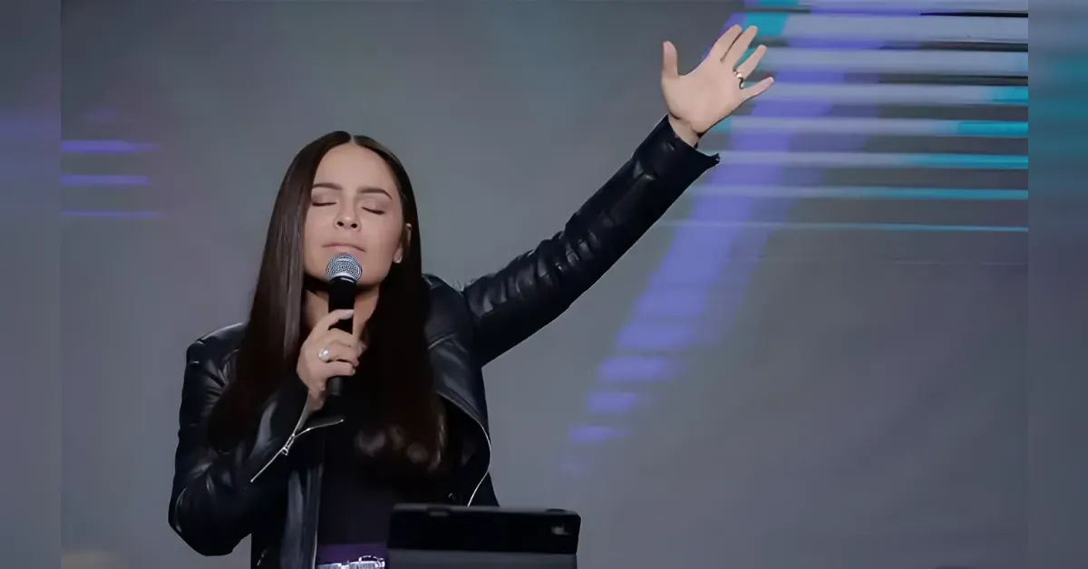 Woman with long dark hair wearing a black leather jacket sings into a microphone with her eyes closed and one arm raised.