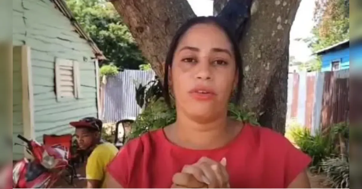 Woman in a red top speaks to the camera outdoors, standing in front of a tree with a man in yellow sitting beside a bike in the background.