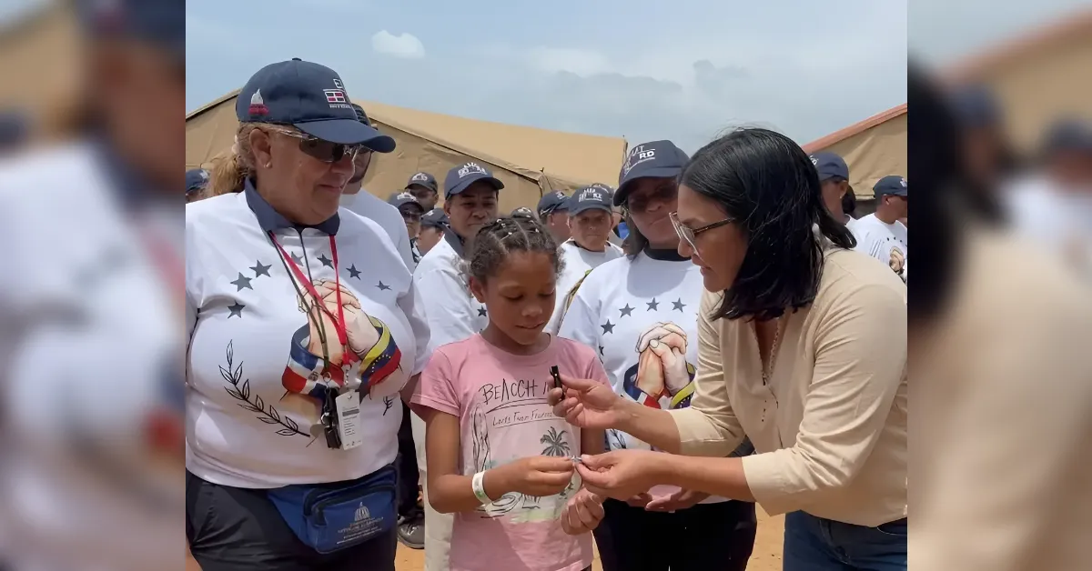 Woman in a beige top hands a small item to a young girl in a pink shirt, as volunteers in white shirts with star designs look on outdoors near tents.