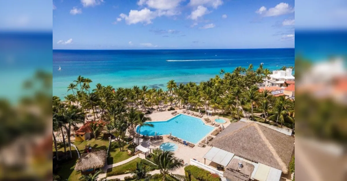 Overhead view of a tropical resort with a large turquoise pool, palm trees, and the blue Caribbean Sea beyond.