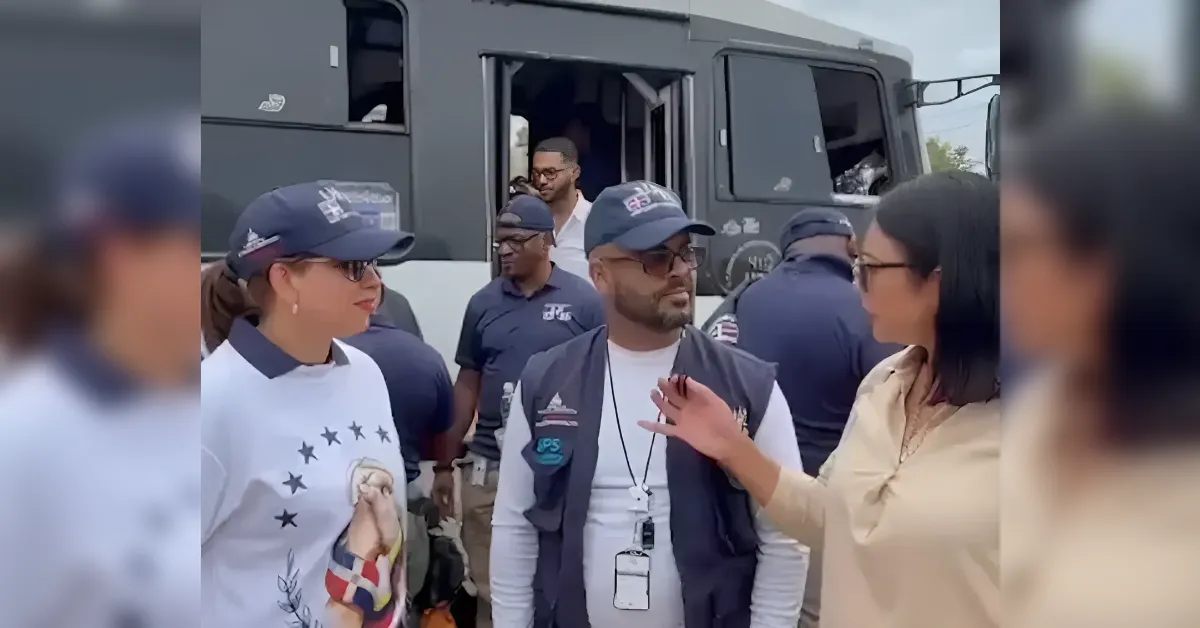Group of people in navy uniforms outside a large black truck; a man with a lanyard and vest speaks to a woman in a beige top.