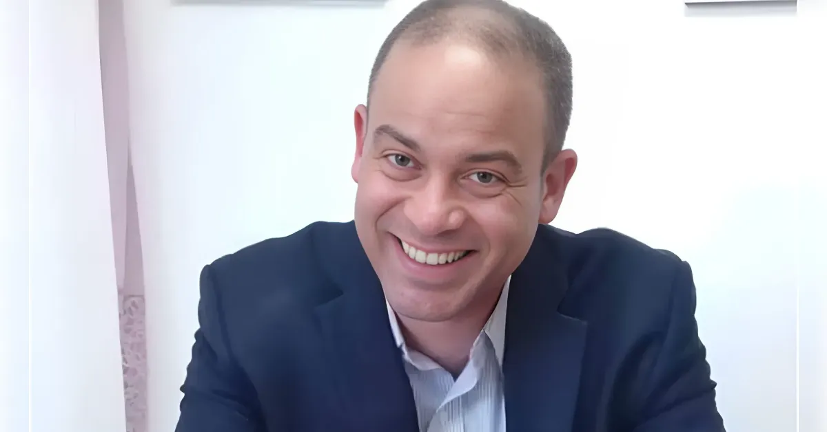 Portrait of a smiling man in a navy blazer and light shirt, seated indoors against a white wall.