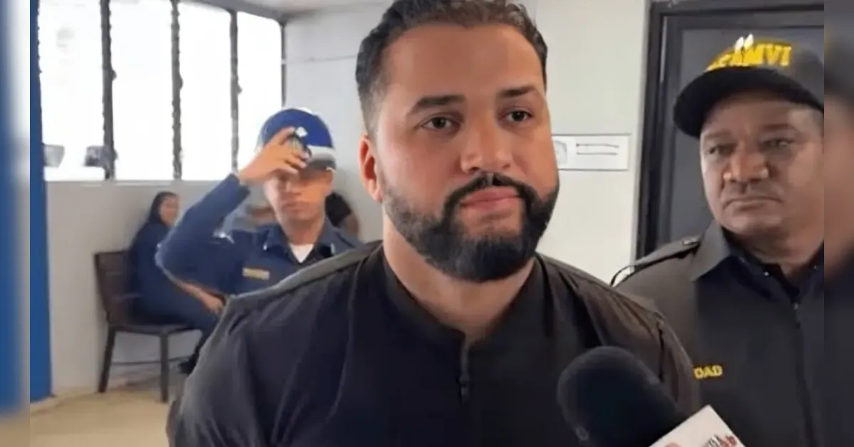 Man with dark beard speaks to a microphone while being interviewed; security guards stand in a hallway behind him.
