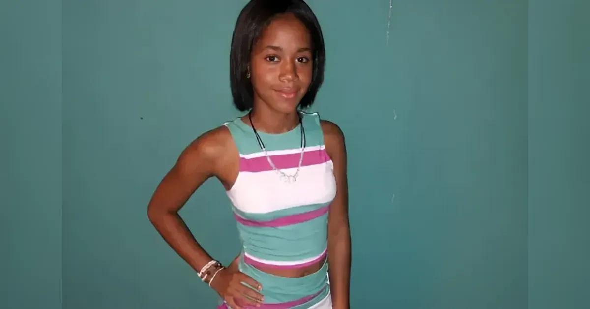 Girl posing with hand on hip, wearing a teal, white, and pink striped sleeveless outfit against a green wall.
