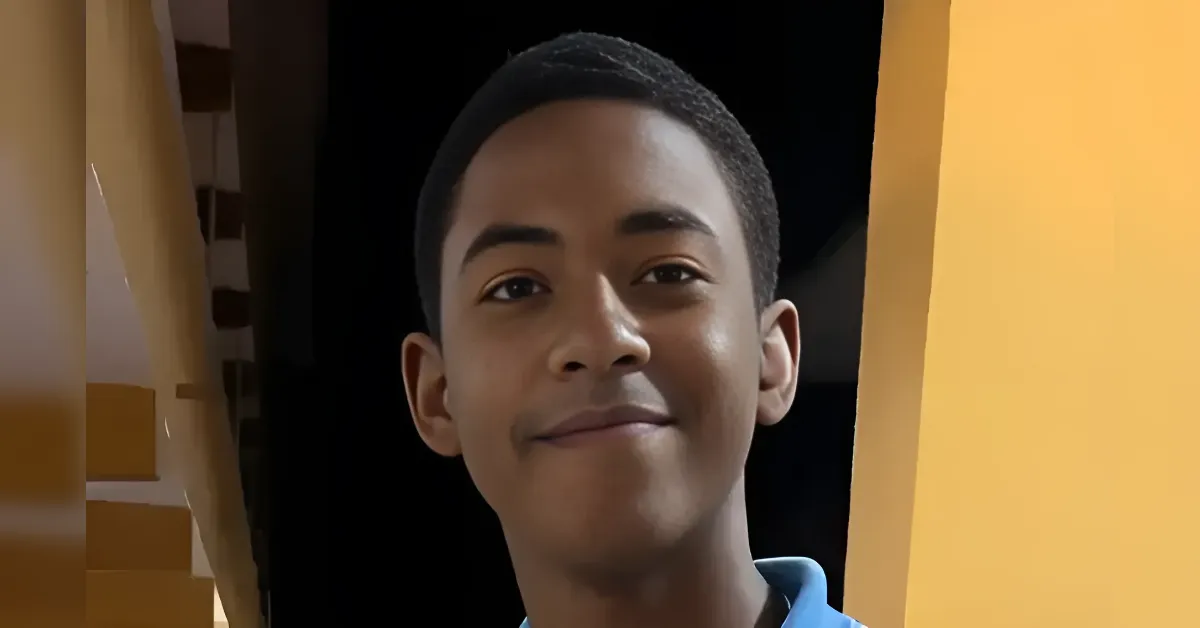 Smiling boy with short dark hair wearing a blue shirt, facing the camera against a dark background.",