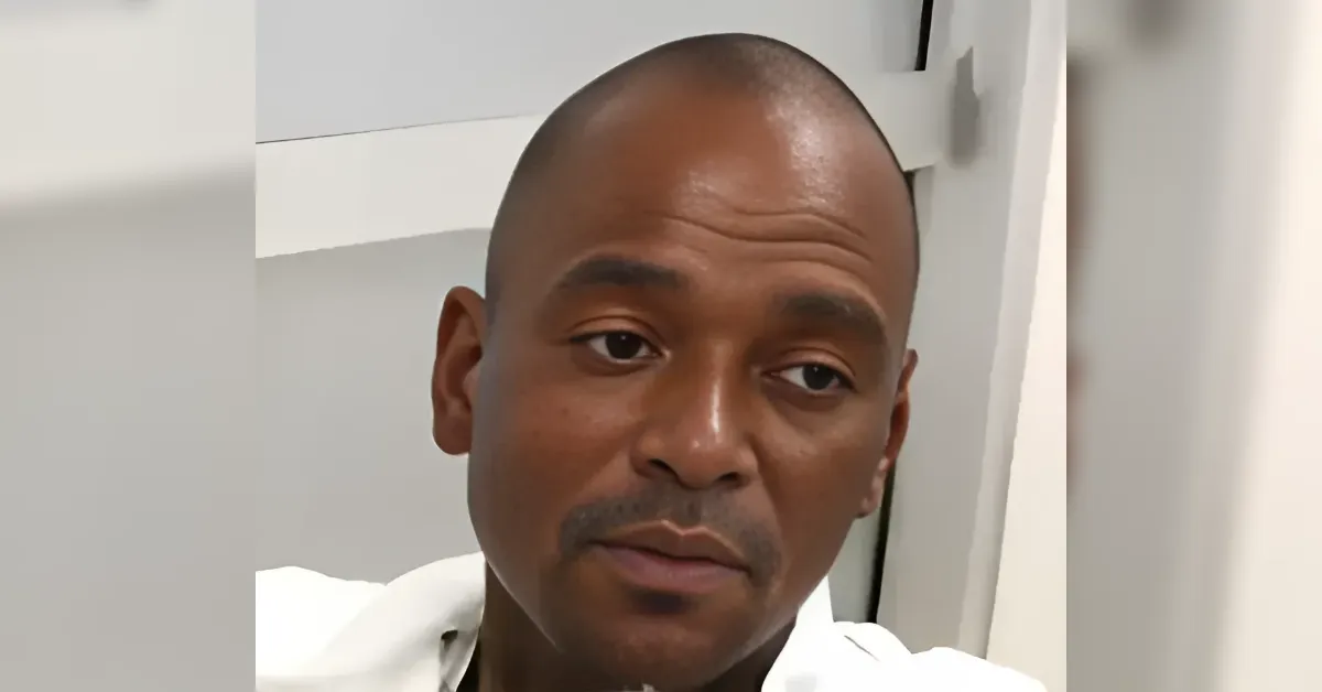 Close-up of a man with a shaved head wearing a white shirt, looking to the side with a tired, thoughtful expression in a neutral room.