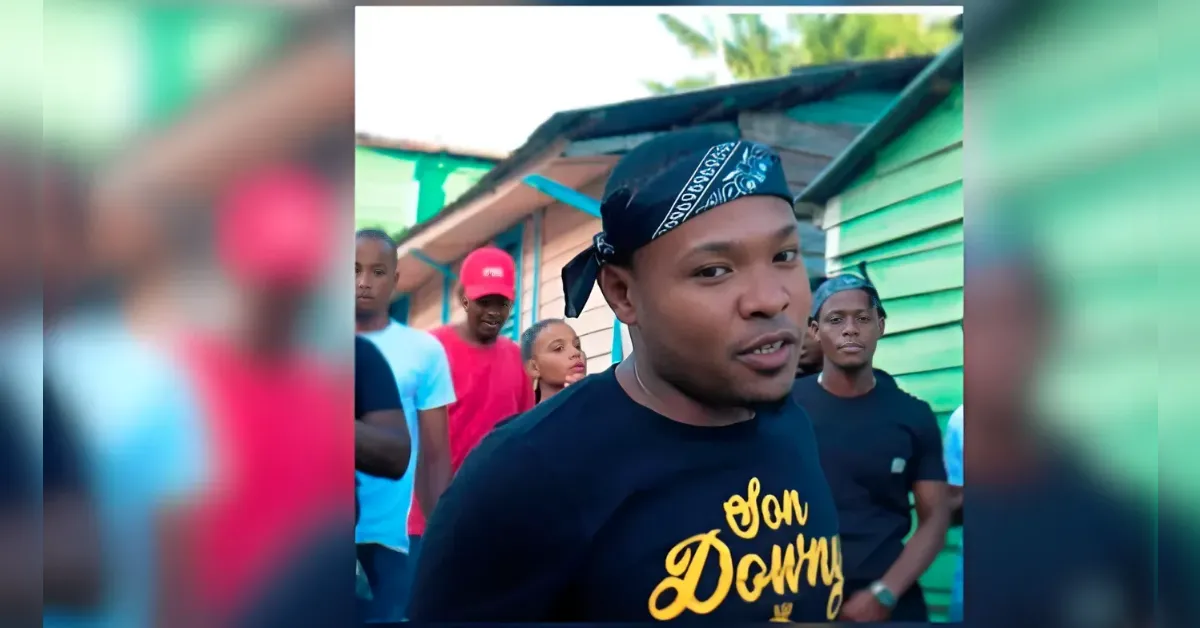 Young man wearing a blue bandana and black shirt with yellow text, speaking to the camera, with friends in a colorful neighborhood behind him.