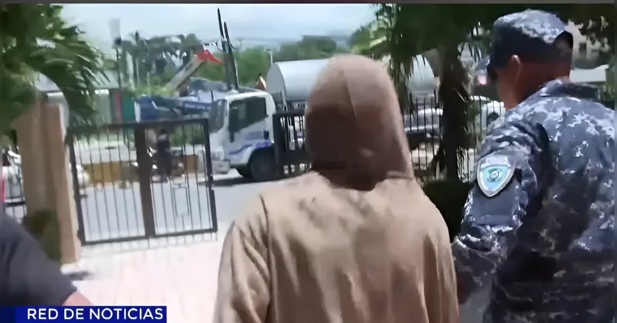 Individual in a tan hoodie walks toward a gated property, with a uniformed officer nearby; a tow truck is visible in the background.