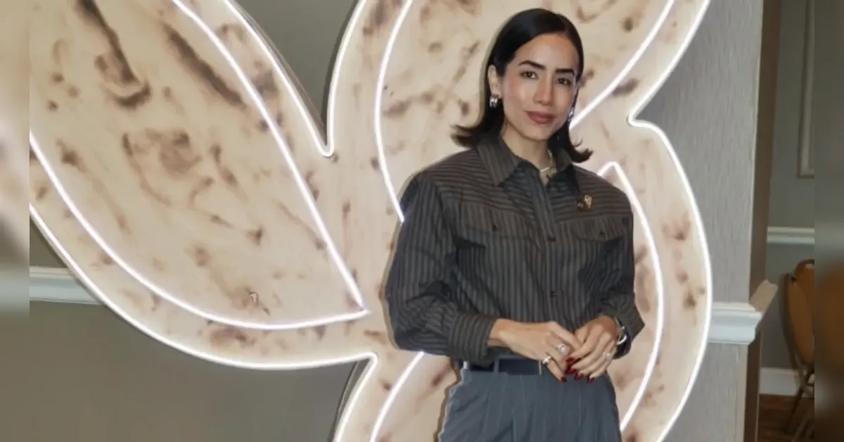 Woman in a striped blouse and gray pants posing in front of a large illuminated decorative sign backdrop.