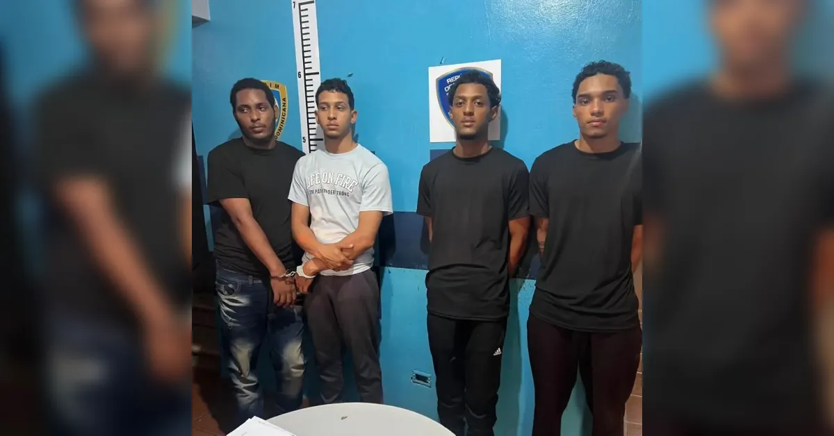 Four young men stand side by side in a mugshot-style lineup against a blue wall with a height chart behind them in what appears to be a police station setting.