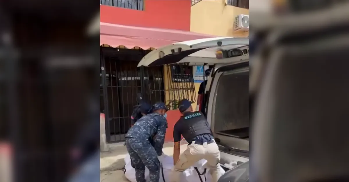 Two uniformed officers lift a white body bag toward an open SUV at the curb in front of a gated storefront.