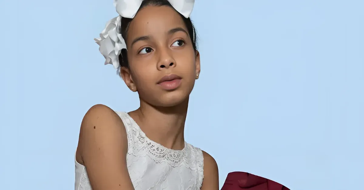 Young girl in a white lace dress with a large white bow in her hair, gazing to the left against a light blue background.