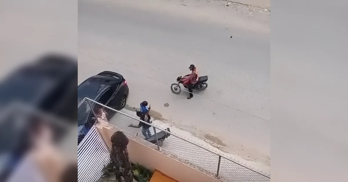 Motorcyclist in a red outfit wearing a helmet riding a motorcycle on a dusty street, pedestrians nearby by a parked car and fence.