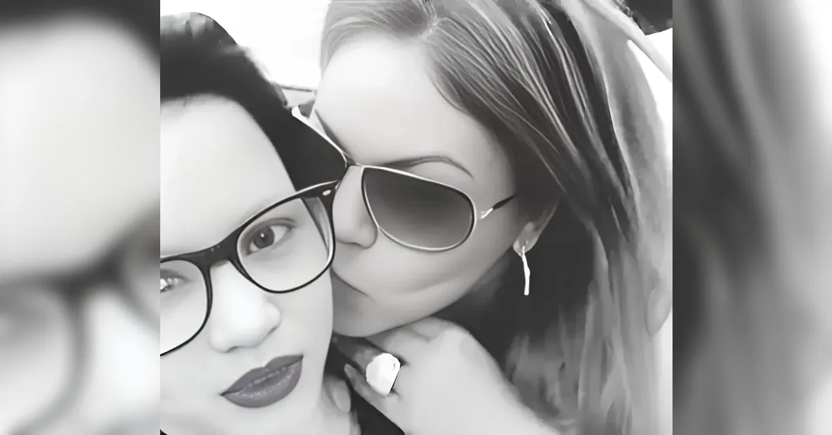 Two women in grayscale share a kiss on the cheek; one wears glasses and bold lipstick, the other sunglasses and earrings.