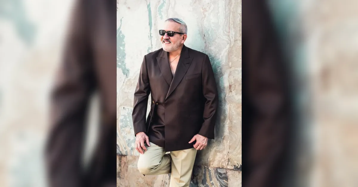 Man in a dark brown blazer and light beige pants leaning against a textured stone wall, wearing sunglasses and smiling outdoors.