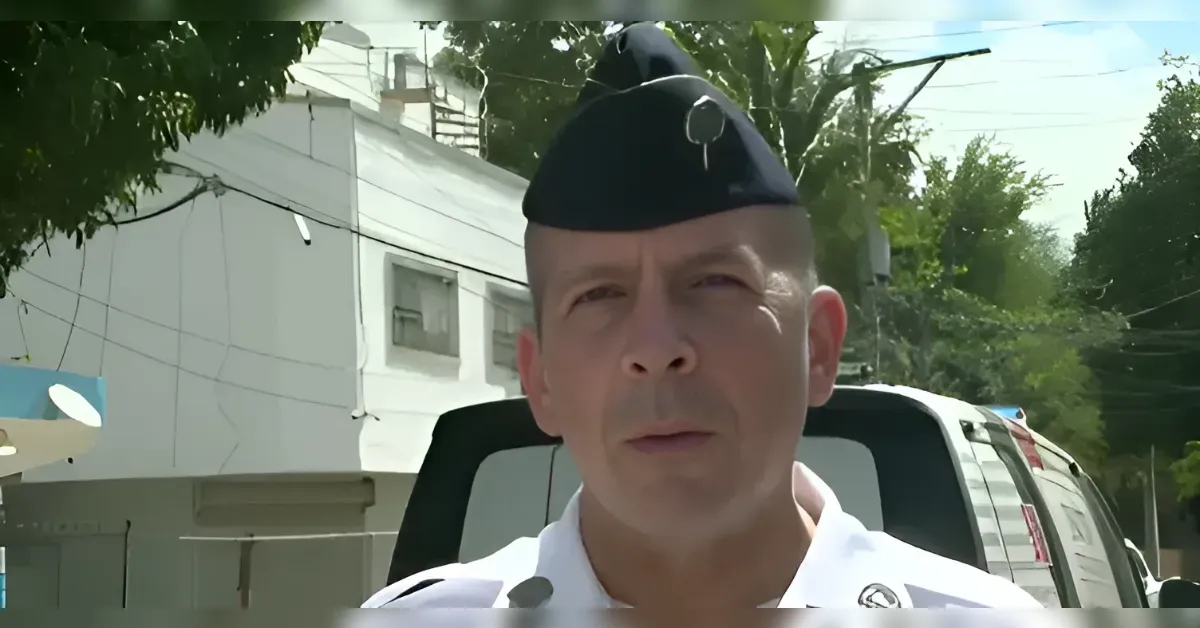 Man in a white uniform and dark military-style cap speaks to the camera on a sunny street with a white van behind him
