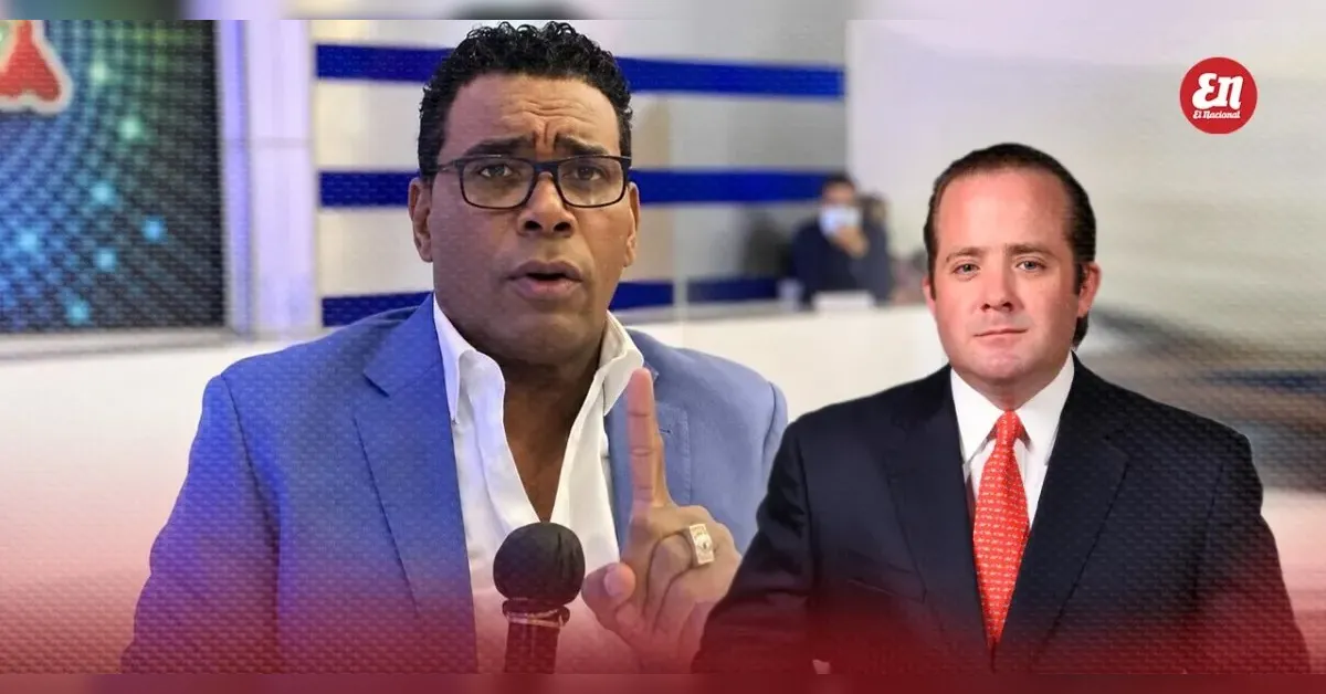 News studio scene with two men; one in a blue suit speaks into a microphone and raises a finger, the other in a dark suit with a red tie beside him.