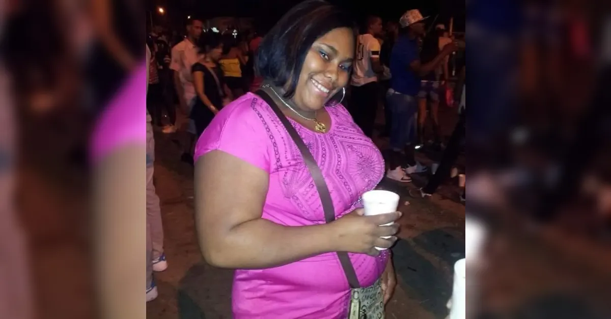 Smiling woman in a pink top holding a white cup at night, with a crossbody bag and a crowd in the background at a street event.