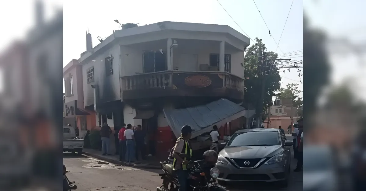 Crowd gathers on a street beside a two‑story corner building with a collapsed awning over the sidewalk; a silver SUV sits nearby on the road.