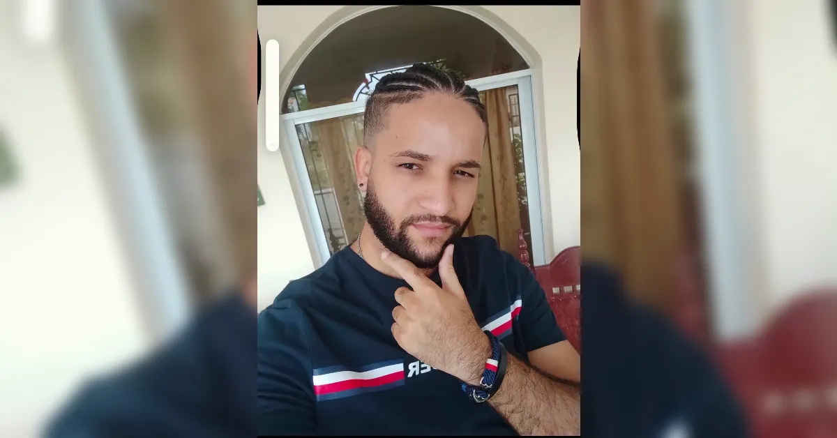 Man taking a selfie on a porch, chin resting on his hand, wearing a black shirt with red and white stripes and a beard.