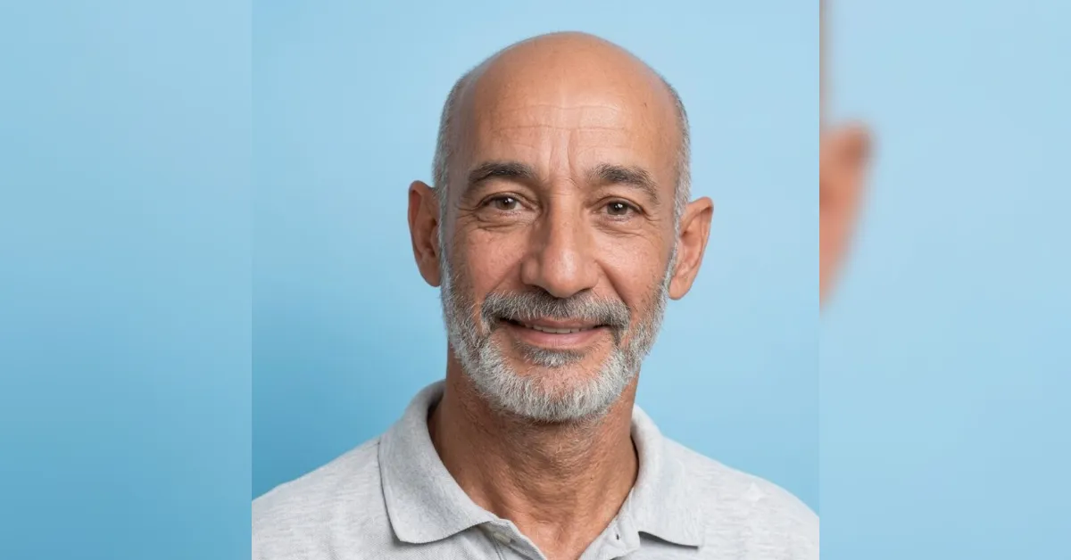 Portrait of a smiling older man with a gray beard and bald head, wearing a light gray polo, against a pale blue background.
