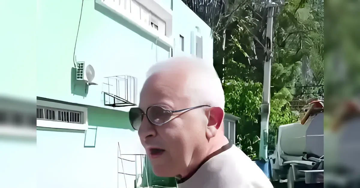 Older man with white hair and sunglasses looking to the side outdoors near a mint-green building.