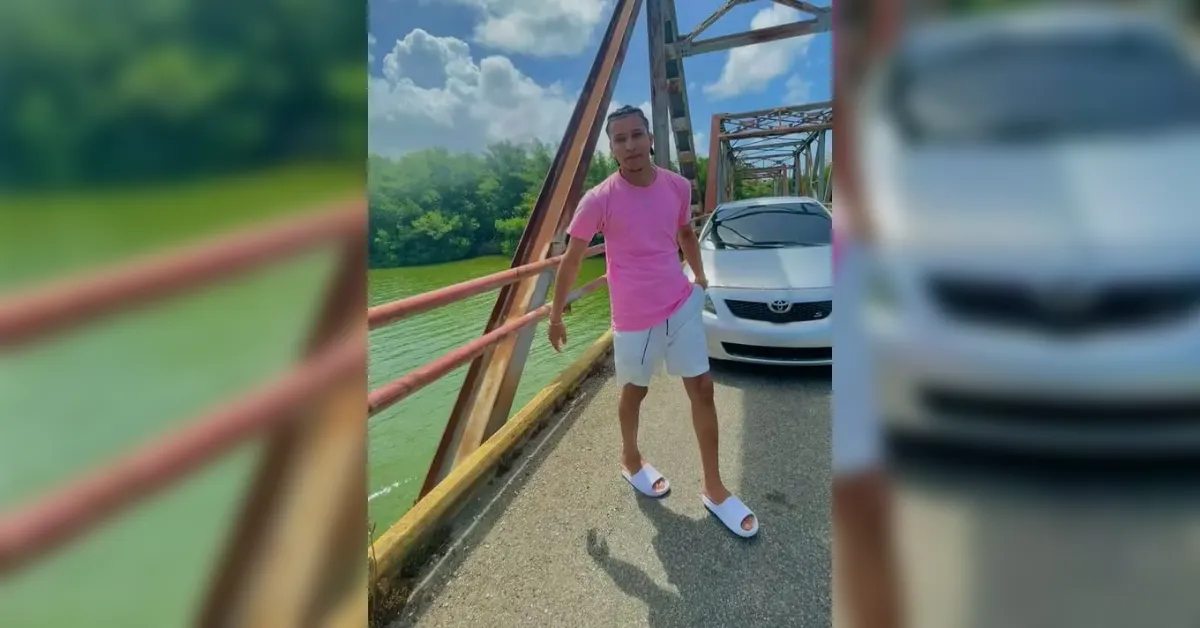 Man in a pink shirt, white shorts, and white slides posing on a bridge beside a white car with a green river and trees in the background.