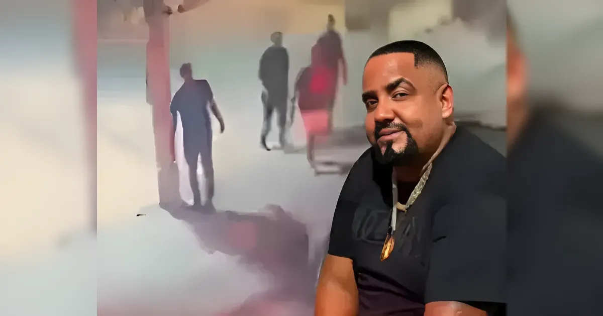 Close-up portrait of a man with a short beard wearing a black shirt and gold necklace, against a blurred, colorful mural background.
