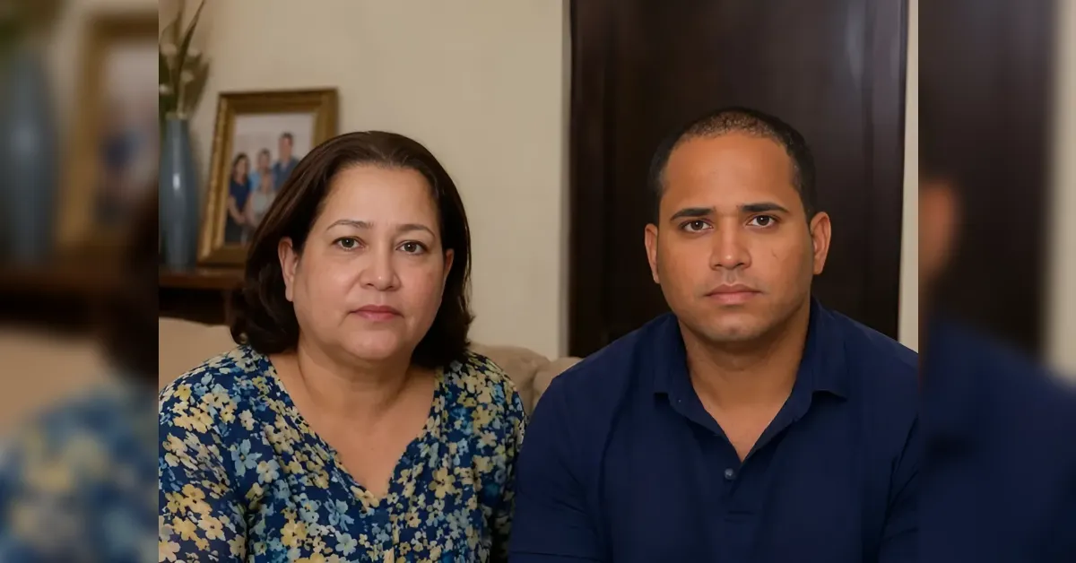 A woman in a blue floral blouse and a man in a dark blue polo sit side by side on a sofa in a home setting, looking at the camera.