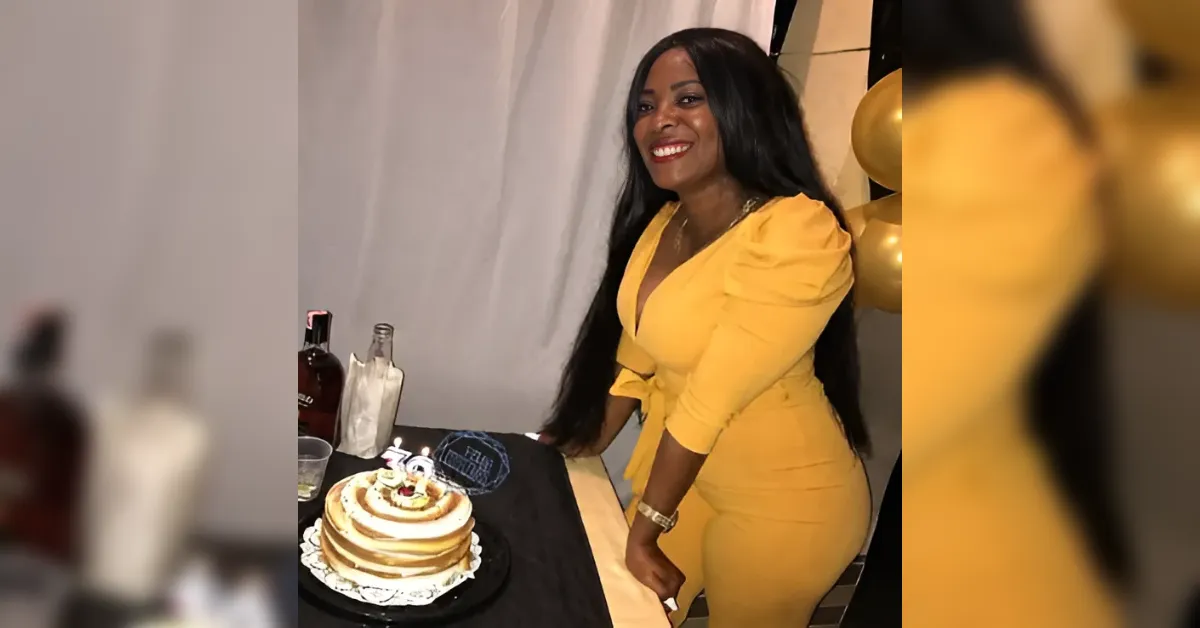 Woman in a yellow dress poses beside a layered birthday cake, smiling at a party.