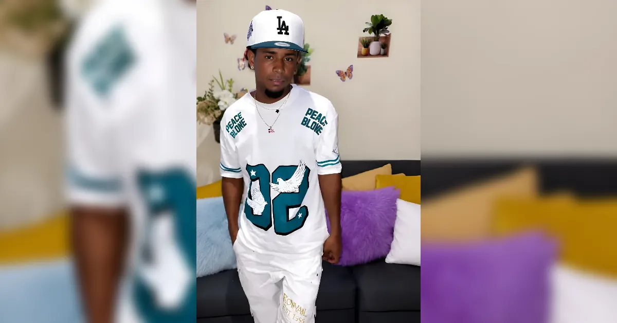Man in a white LA cap and jersey with teal 92 and dove graphics poses in a living room with purple and yellow cushions on a black sofa.