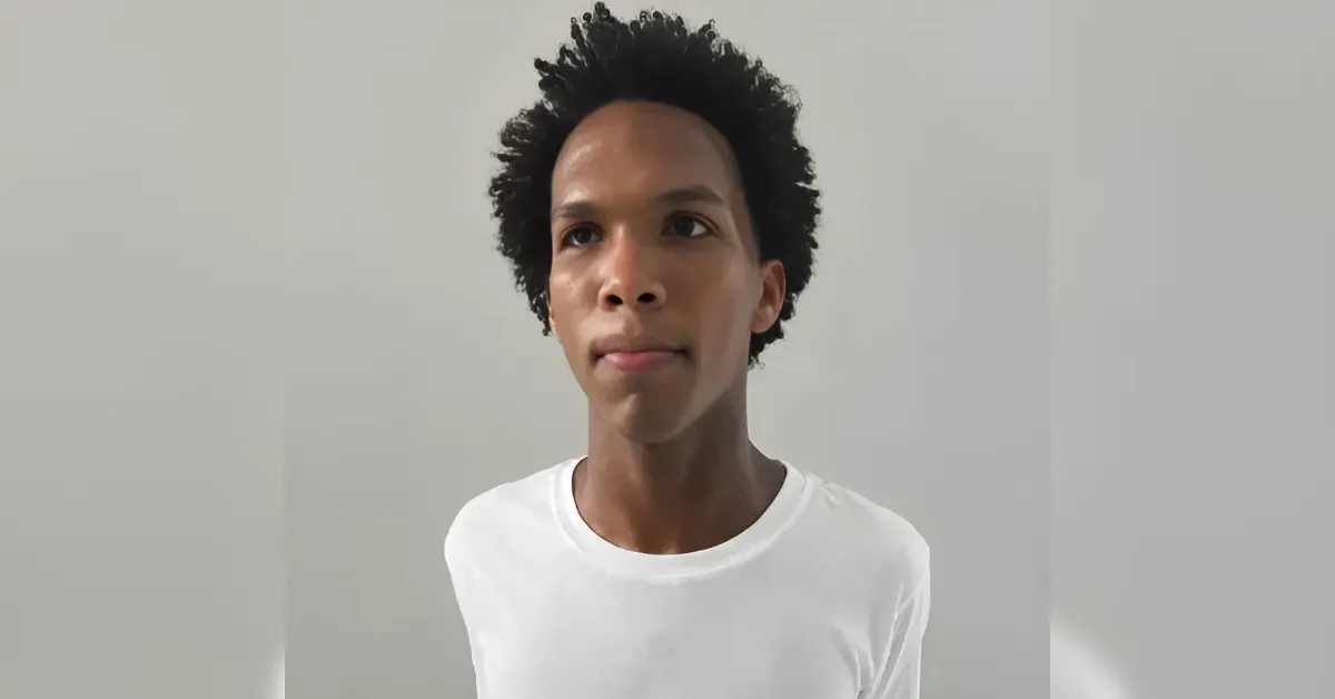 Young person with short curly hair wearing a white T-shirt, standing against a light gray wall.