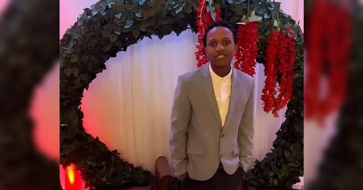 Young man in a light gray blazer and white shirt standing in front of a circular leafy arch with red hanging flowers at a decorated event backdrop.