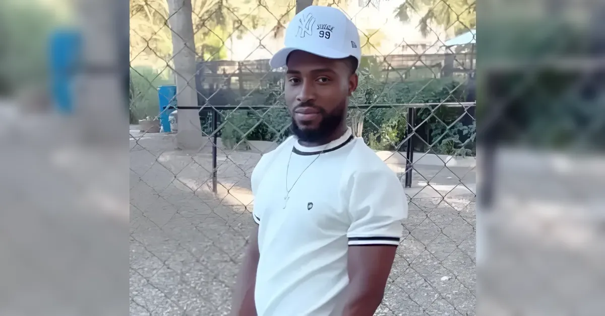 Young man in a white polo shirt and white cap posing outdoors in front of a chain-link fence.