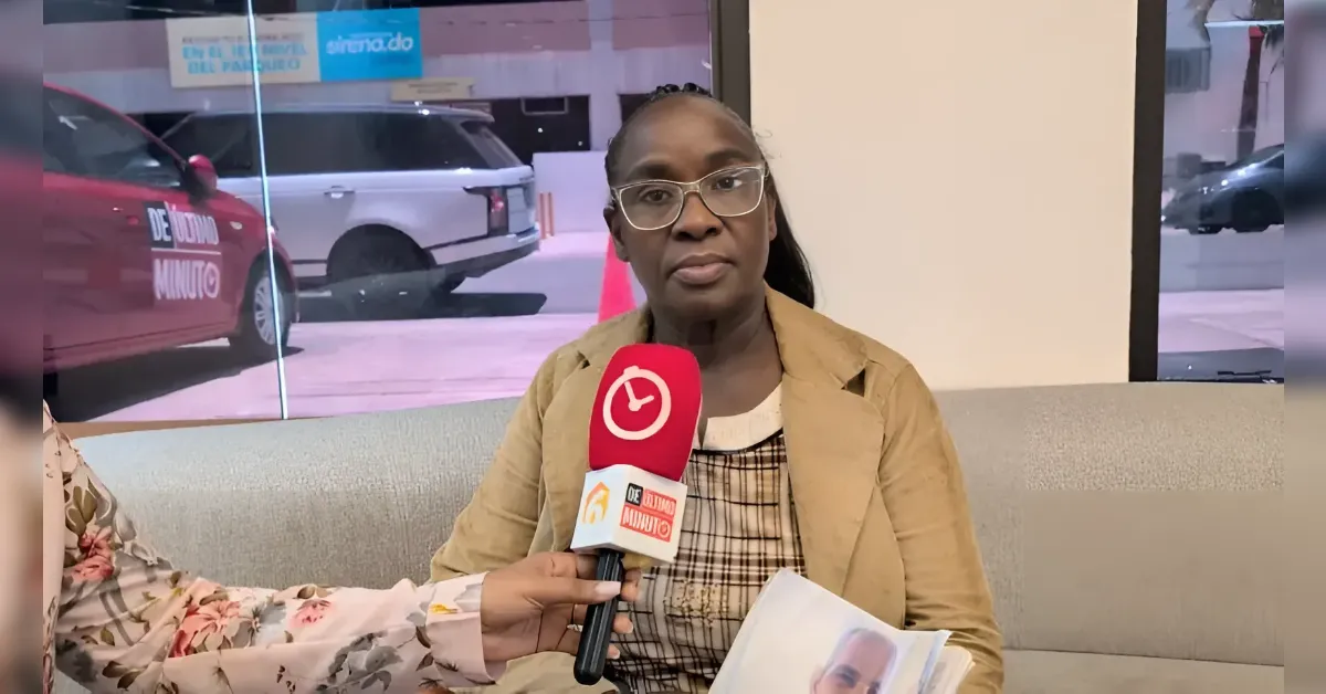 Woman with glasses in a tan blazer sits on a couch during an interview, holding a magazine, microphone held near her face by an interviewer.