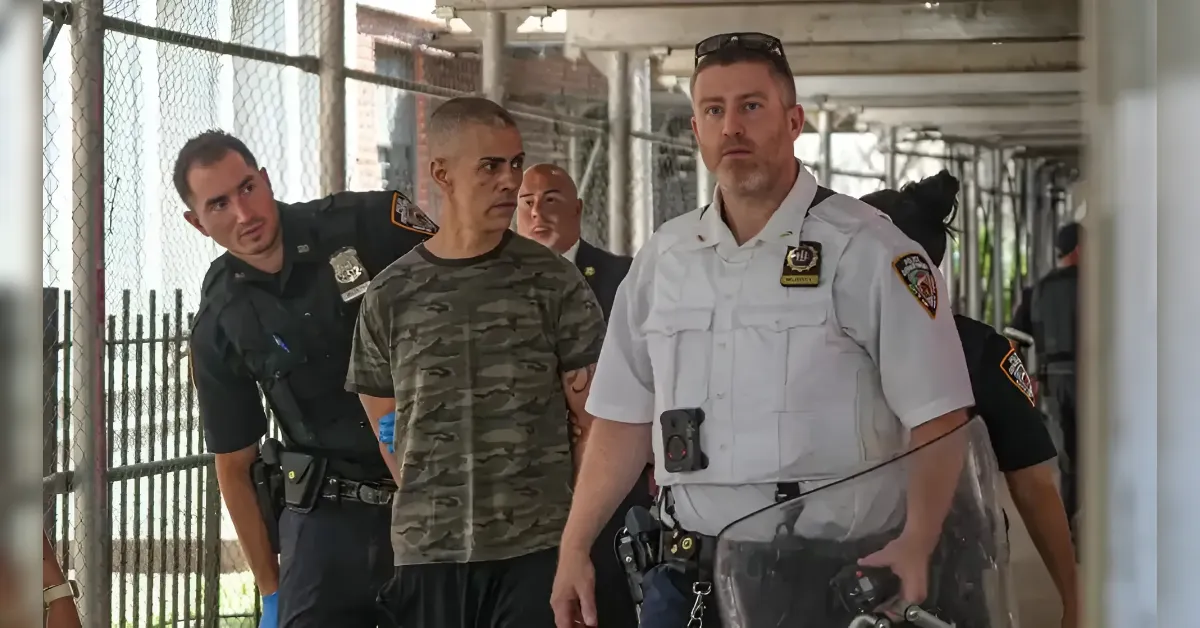 Group of uniformed officers escorting a man in camouflage shirt down a fenced corridor.