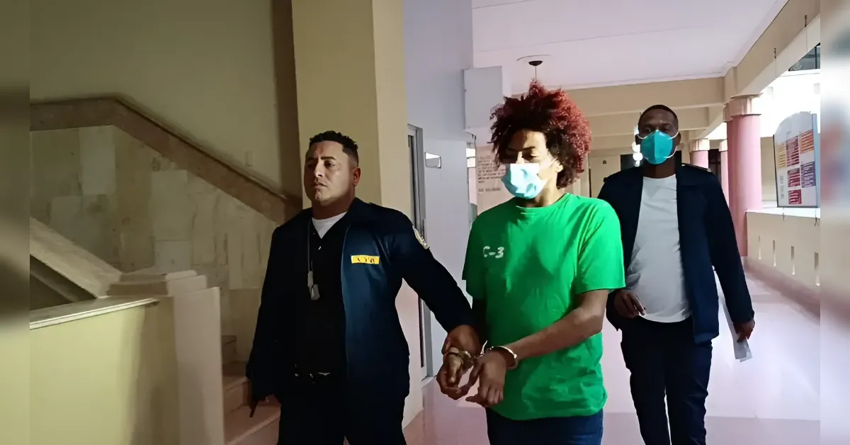 Detainee in a green shirt in handcuffs is escorted by two men down a hallway, both wearing masks.
