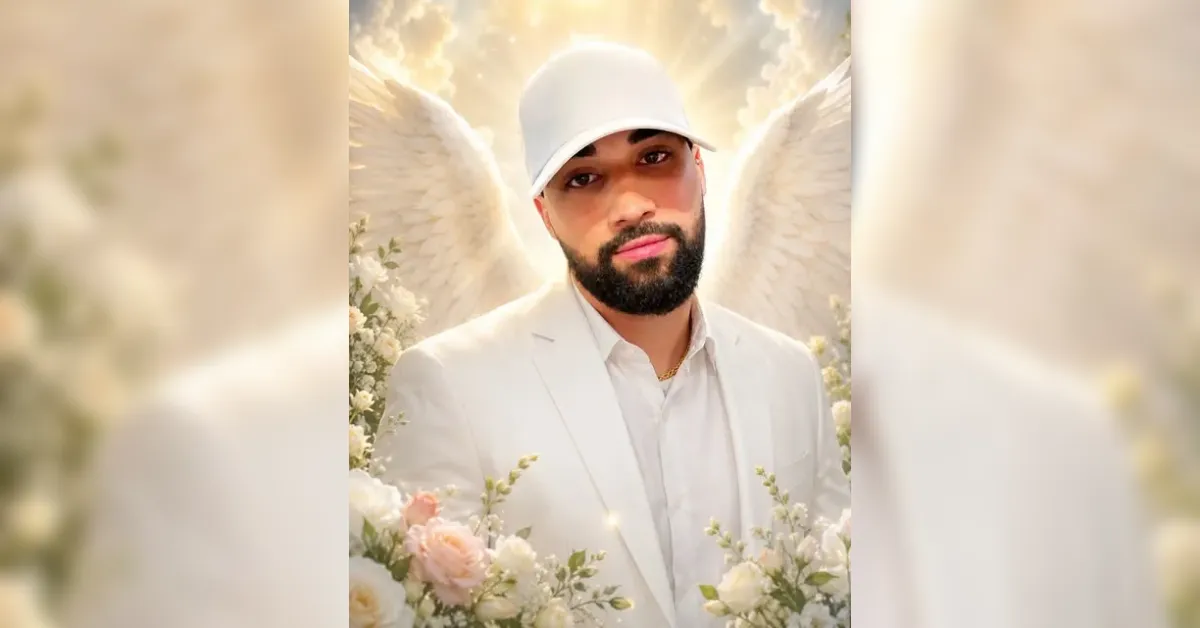 Man with a beard in a white suit and cap, angel wings behind him among pale flowers.