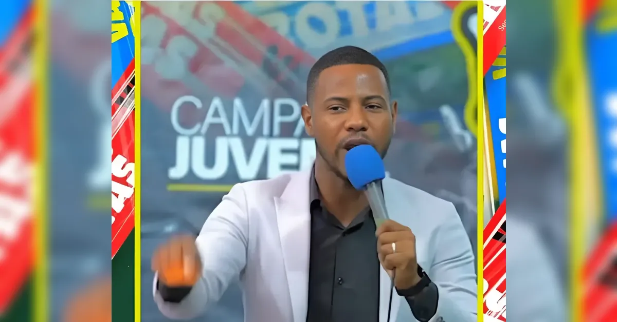 Male speaker wearing a light blazer speaks into a blue microphone on stage, gesturing toward the audience with a blurred backdrop that reads 'CAMP JUVEN'.