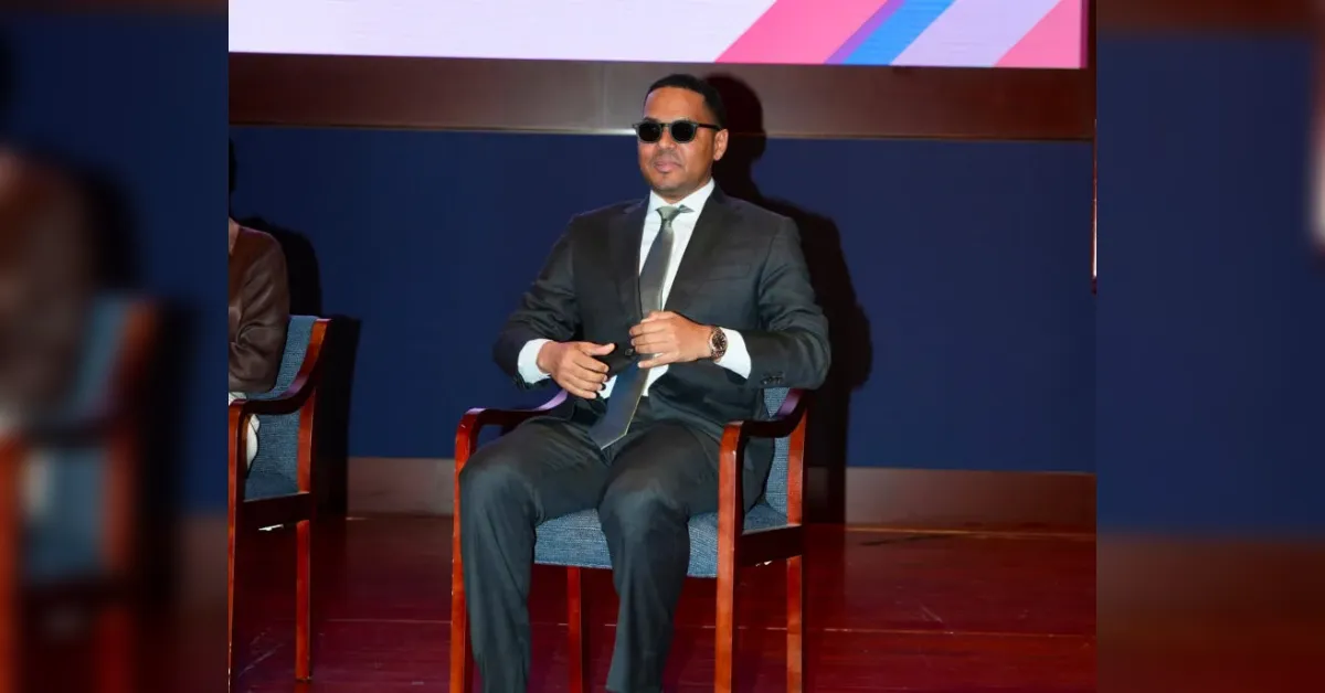 Man in a dark suit and sunglasses sits on stage in a chair, adjusting his tie against a blue backdrop.