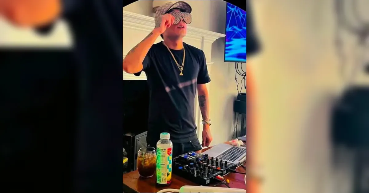 DJ wearing a patterned mask and cap, adjusting his mask while standing at a laptop and mixer.